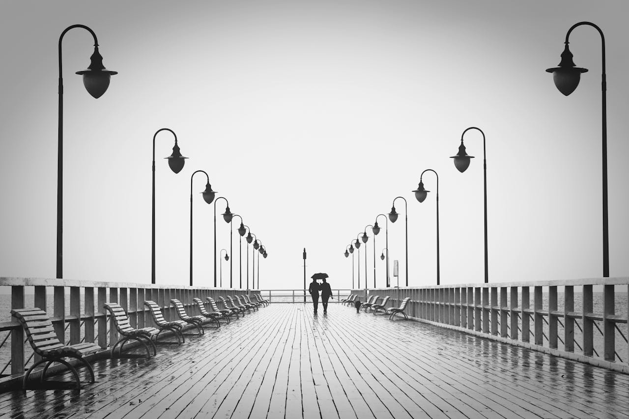 about-img-01 A couple holding umbrellas walks on a rainy boardwalk, embodying romance and tranquility.